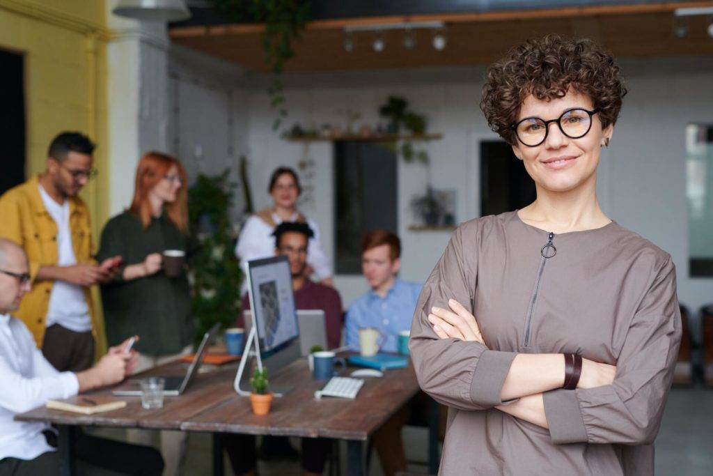 Smiling woman facing camera while her coworkers are in a meeting at the table behind her